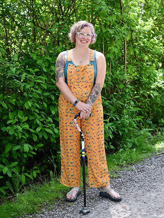 "A woman with shirt, curly purple hair and wearing glasses is smiling outside, standing with a can. She is wearing yellow overalls with a print on them and she has tattoos on her arms."