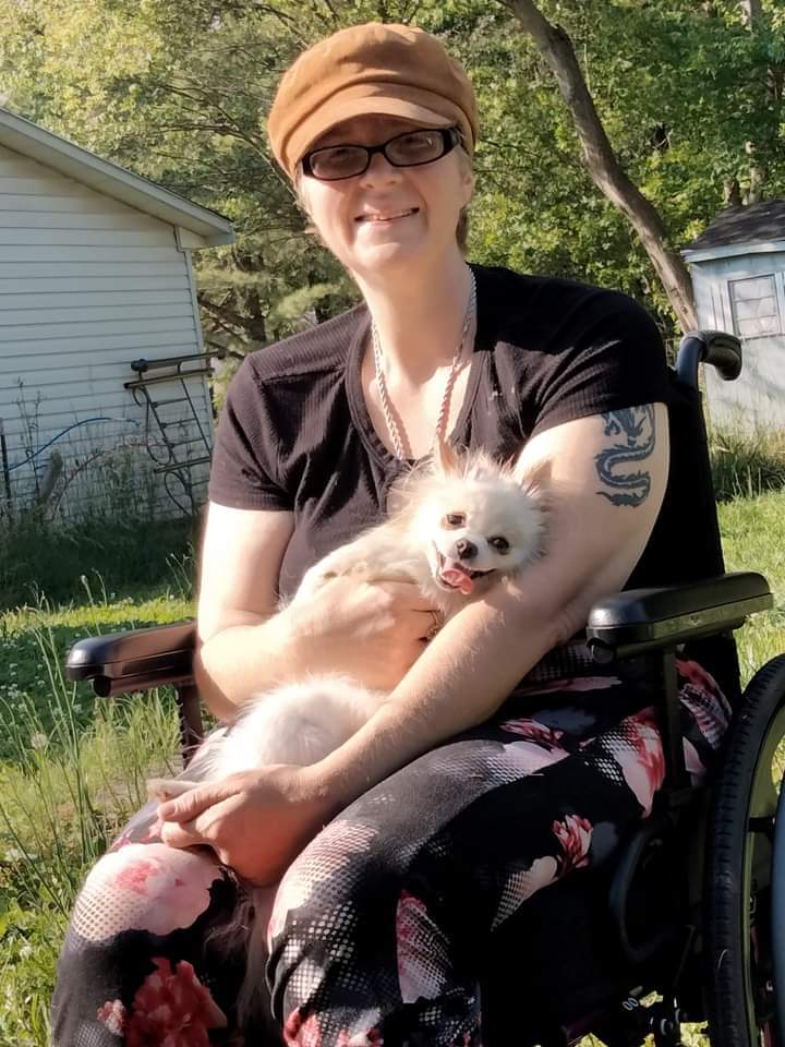 A white woman wearing black framed glasses, a black shirt, and a tan cap is sitting in a wheelchair holding a small dog. The dog has its tongue out. A tattoo is visible on the person's left arm.