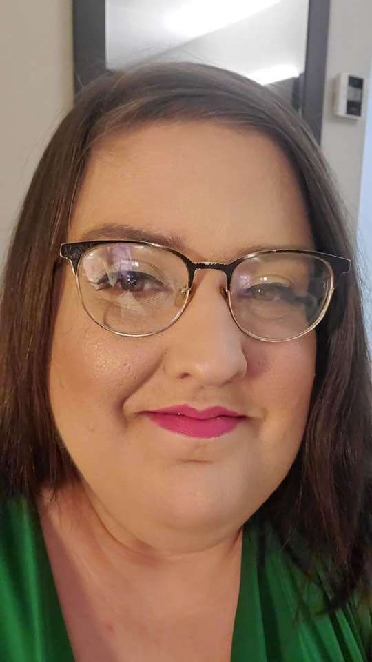 A white woman with brown hair, stylish glasses, and bright lipstick smiling. She is wearing a green shirt.