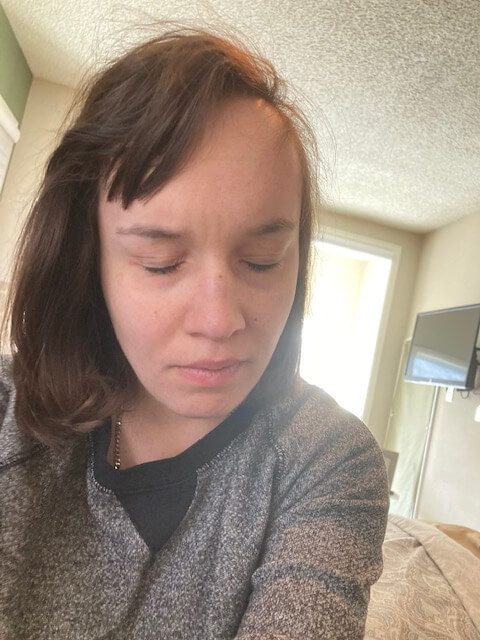 A white woman with brown shoulder-length hair and brown bangs to the side. She has her eyes closed and is wearing a gray shirt.