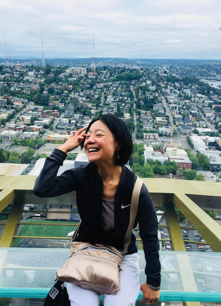 An Asian woman with short, dark hair is laughing and smiling. She has her hand up to her face and is sitting on the top of the Space Needle in Seattle with an expansive view behind her. She is wearing a black top and a purse that is sitting on her lap.
