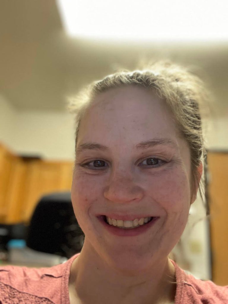 A white woman with brown hair that is pulled back is smiling. She is wearing a pink shirt.
