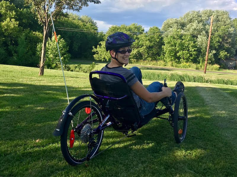 A white woman is wearing a helmet and sunglasses and riding a trike bicycle. She is looking back at the camera over her shoulder and smiling. It is a bright sunny day and she is on green grass with trees in the background.