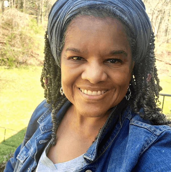 A black woman wearing a grey headband and silver earrings is smiling at the camera. She has grey hair styled in long thin locs and is wearing a blue jean jacket. There is green grass and sunshine in the background.