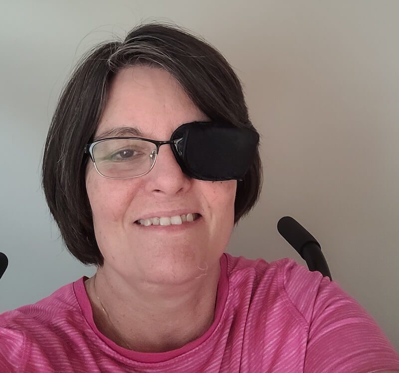 A white woman with short brown hair is smiling at the camera. She is wearing glasses with a black eye patch over one eye, and a pink shirt.