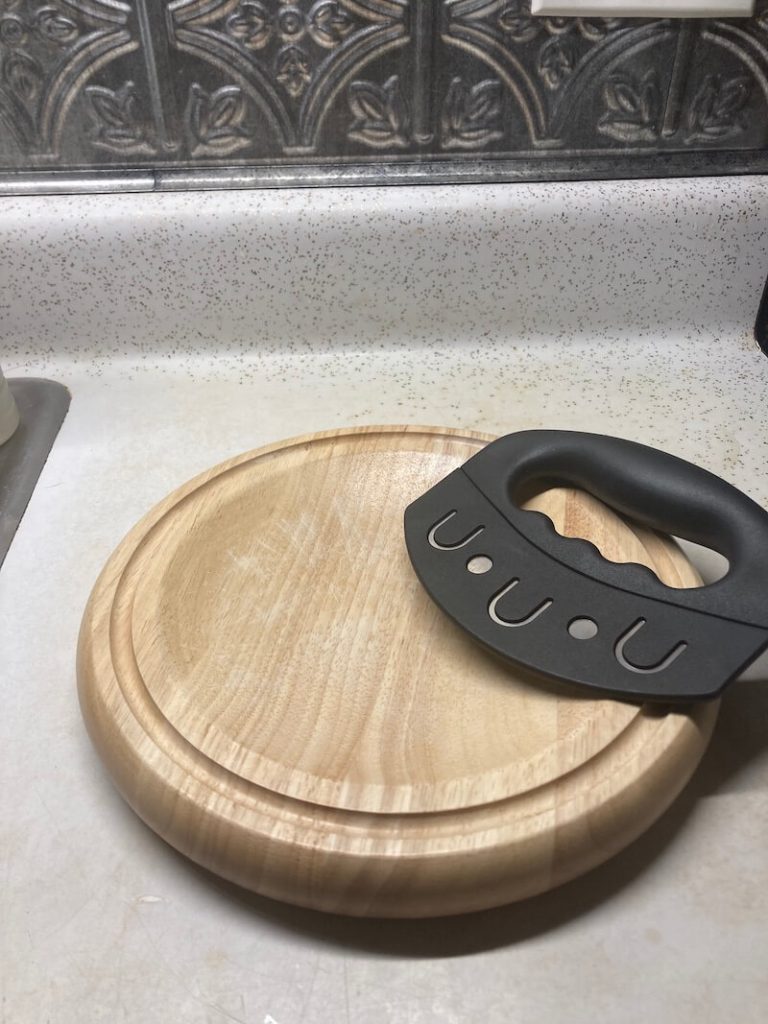 A mezzaluna knife with a black handle is sitting on a wood, circular cutting board. The blade of the knife is covered with a black sheath.