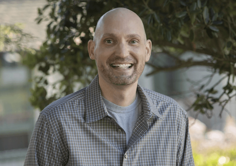 A white man with no hair and a cropped beard smiles at the camera wearing a button down collared shirt in front of a tree background.