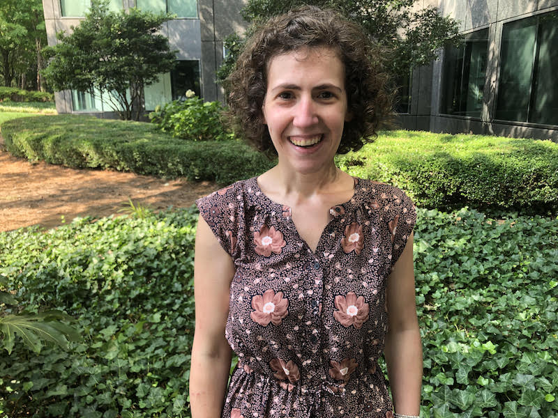A white woman with short brown curly hair is smiling in front of green bushes on a sunny day. She is wearing a dress with pink flowers.