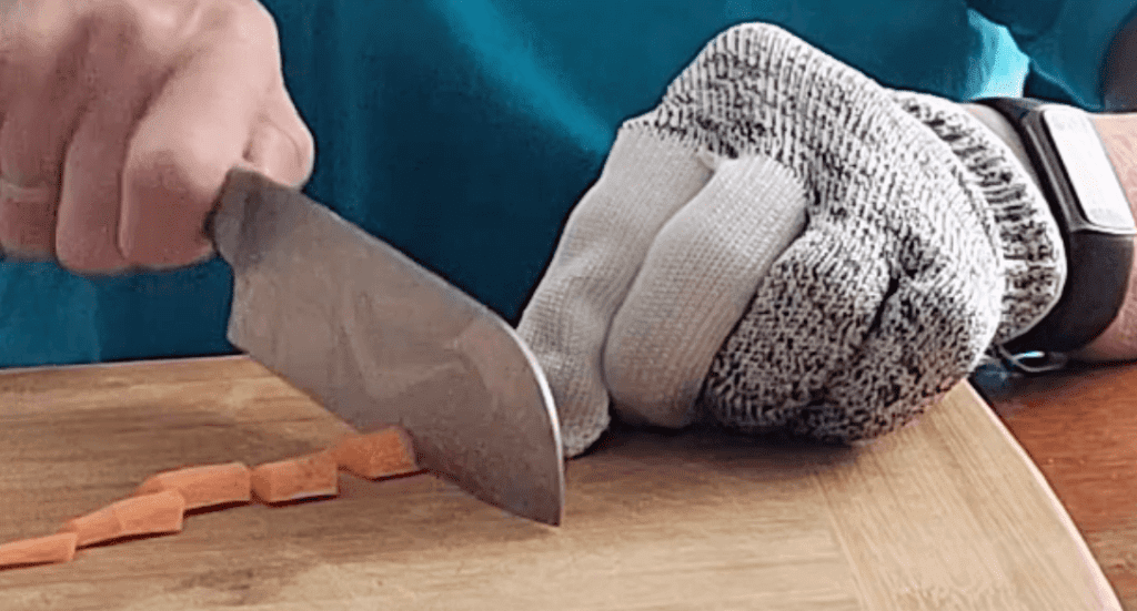 A person is using a large knife to chop a carrot on a cutting board. The hand that is holding the carrot (not holding the knife) has a gray and white fabric (of some type) glove on it.