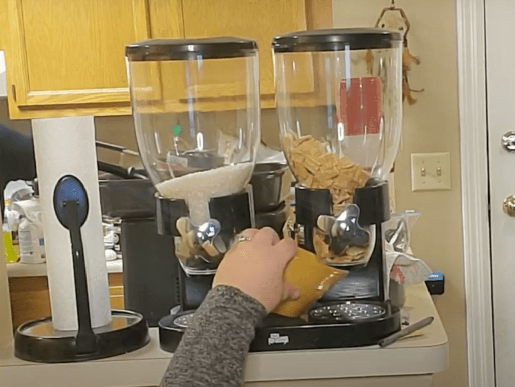 Two large clear dispensers with black and silver accents sitting on a kitchen countertop. A hand reaches up to it with a yellow bowl. One side is filled with rice and the other with cereal. Each container has a silver knob in the shape of an x, which when turned dispenses the food.
