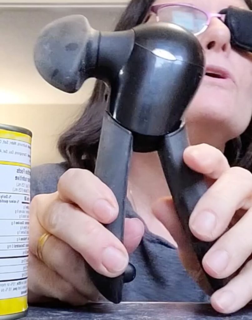 A person is holding a black OXO Good Grips Smooth Edge Can Opener by its handles, pulling them slightly apart. The large turning knob is visible on the left side.