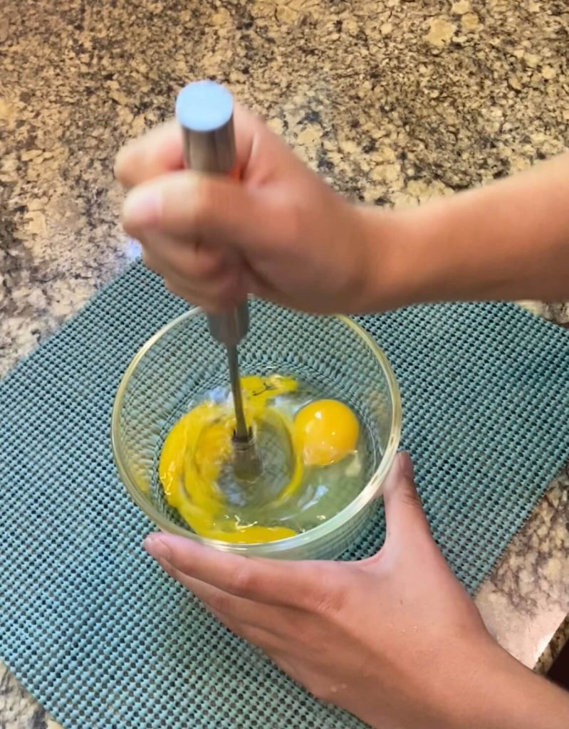 Whisk in action, being pushed up and down, to whisk eggs. The bowl is sitting on a teal anti-slip placemat