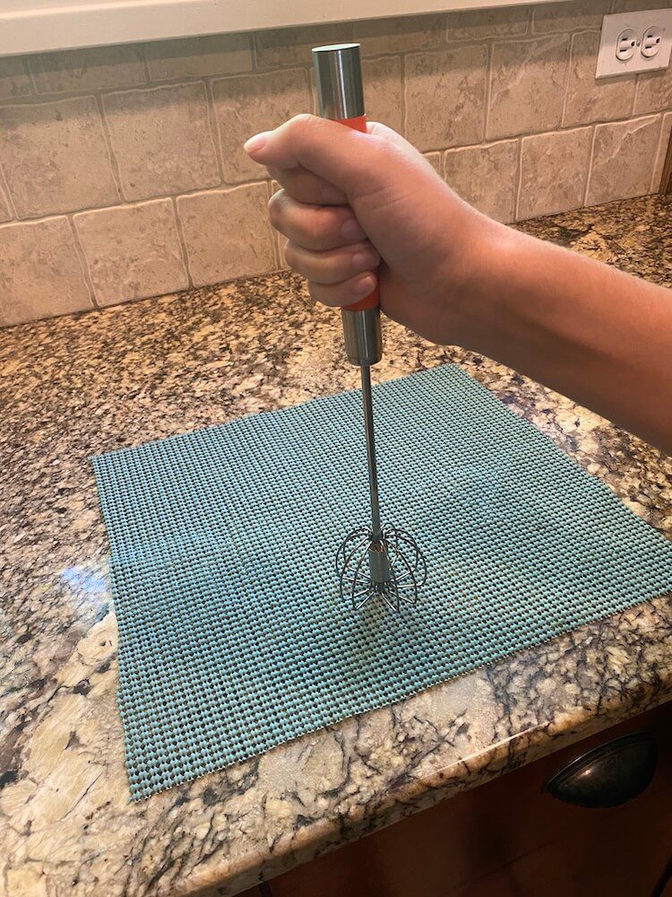 The metal whisk with a red handle, being held on a kitchen counter