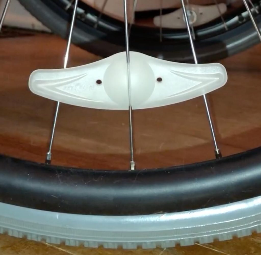 Spoke light is attached to spoke on a wheelchair wheel. It is not lit up and looks like small plastic wings, going across 3 spokes.