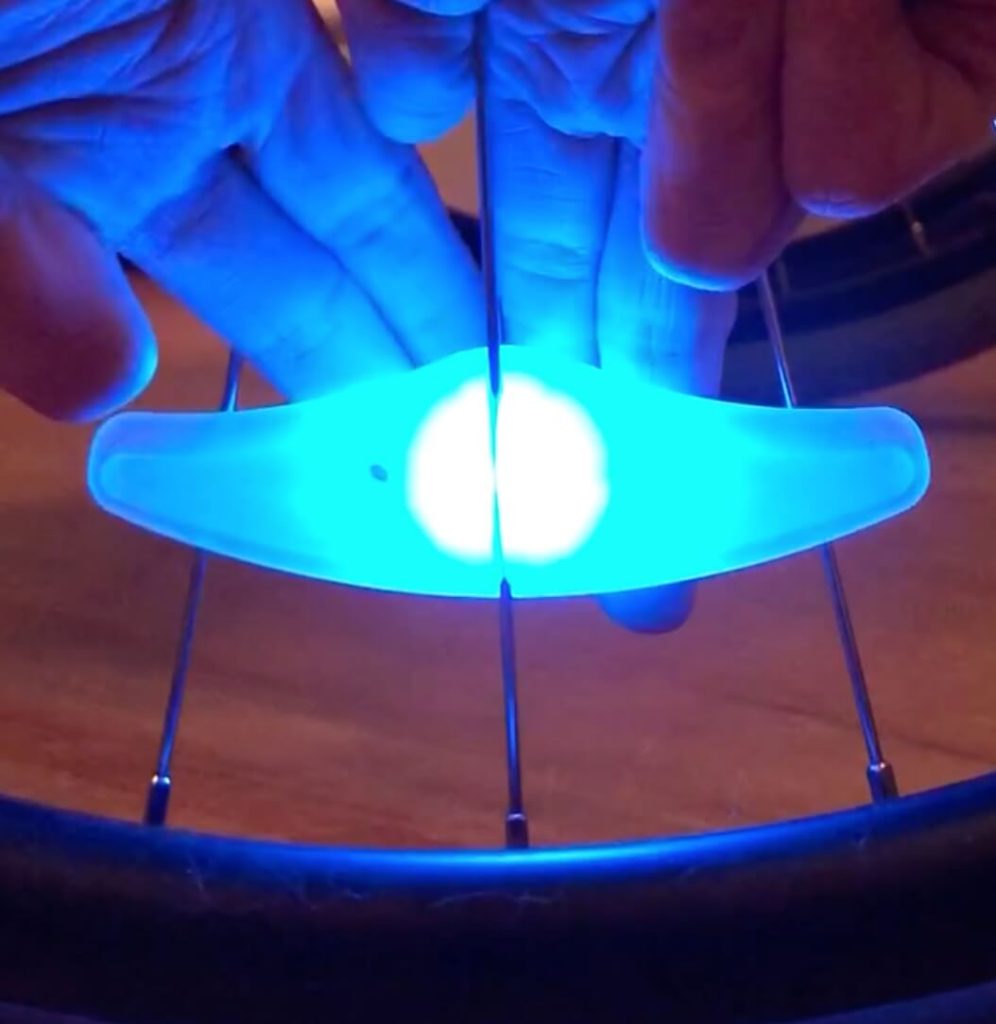 Spoke light lit up in blue. It is attached to the spokes of a wheel and a person is pushing a button on the back using several fingers.