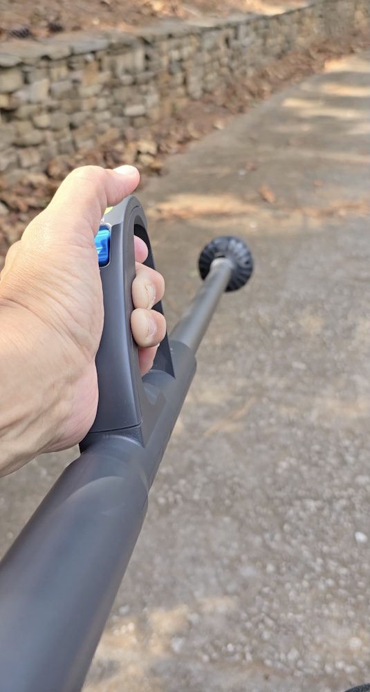A person is holding the handle of a thin, gray stick blower. There is a blue button visible beneath their thumb, and we see the whole way down the device to the blowing end.