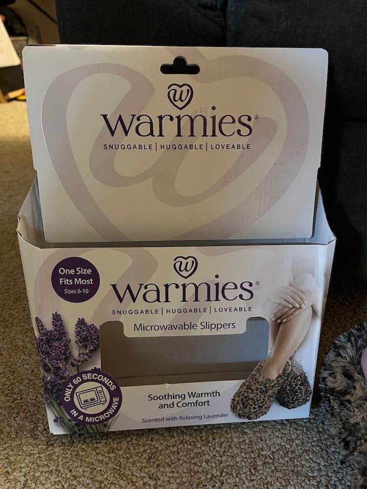 Front of the warmies microwavable slippers box with a picture of the leopard slippers. Says "One size fits all" and "soothing warmth and comfort - Scented with Relaxing Lavender".