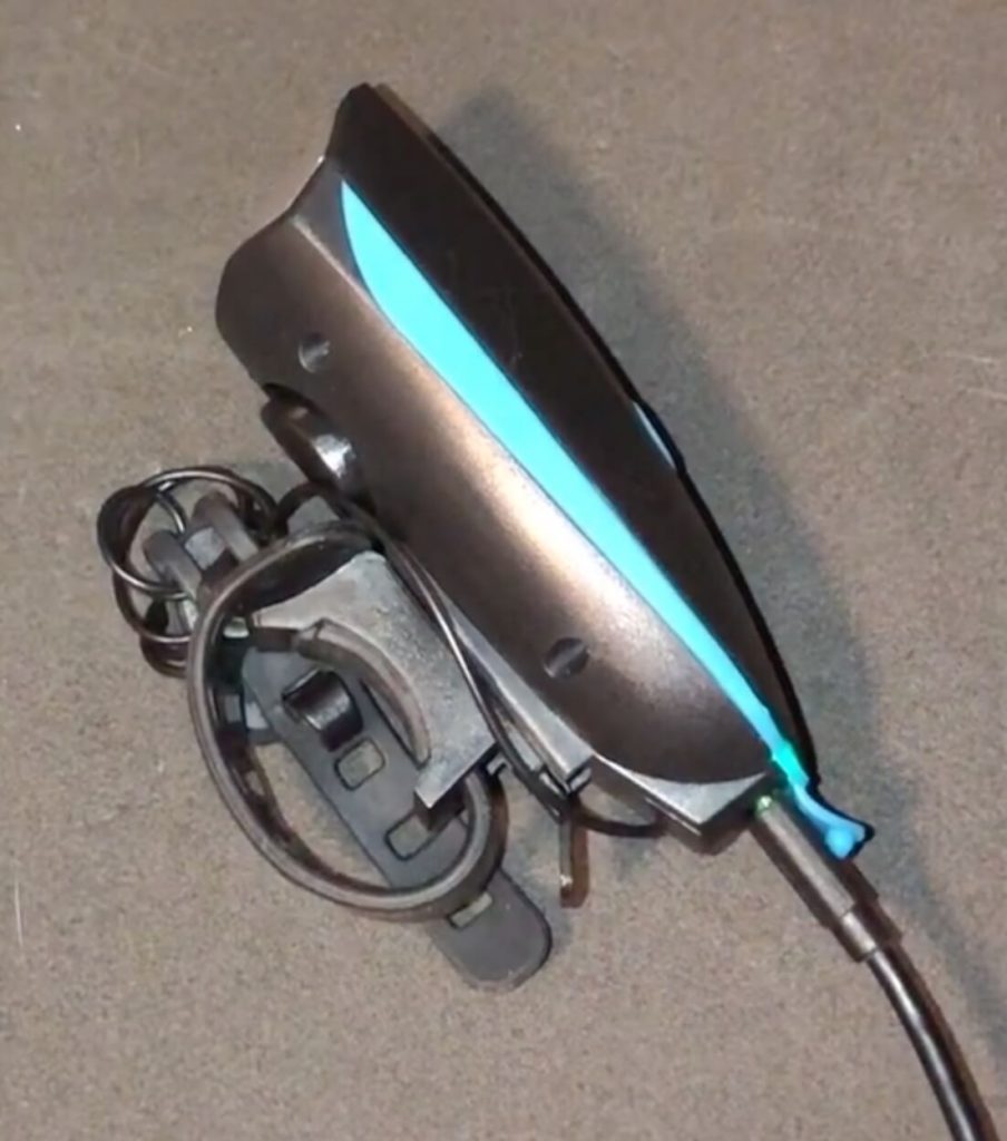 Side view of front light plugged into charging cable. Device is black with light blue accent and has a round loop strap with notches for attaching to device.