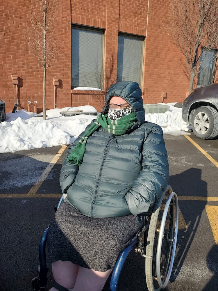 Ericka bundled up in her dark green (called "darkest spruce" by the manufacturer) down jacket with a scarf wrapped around her face. She is in her wheelchair in a parking lot. There is snow in the background.