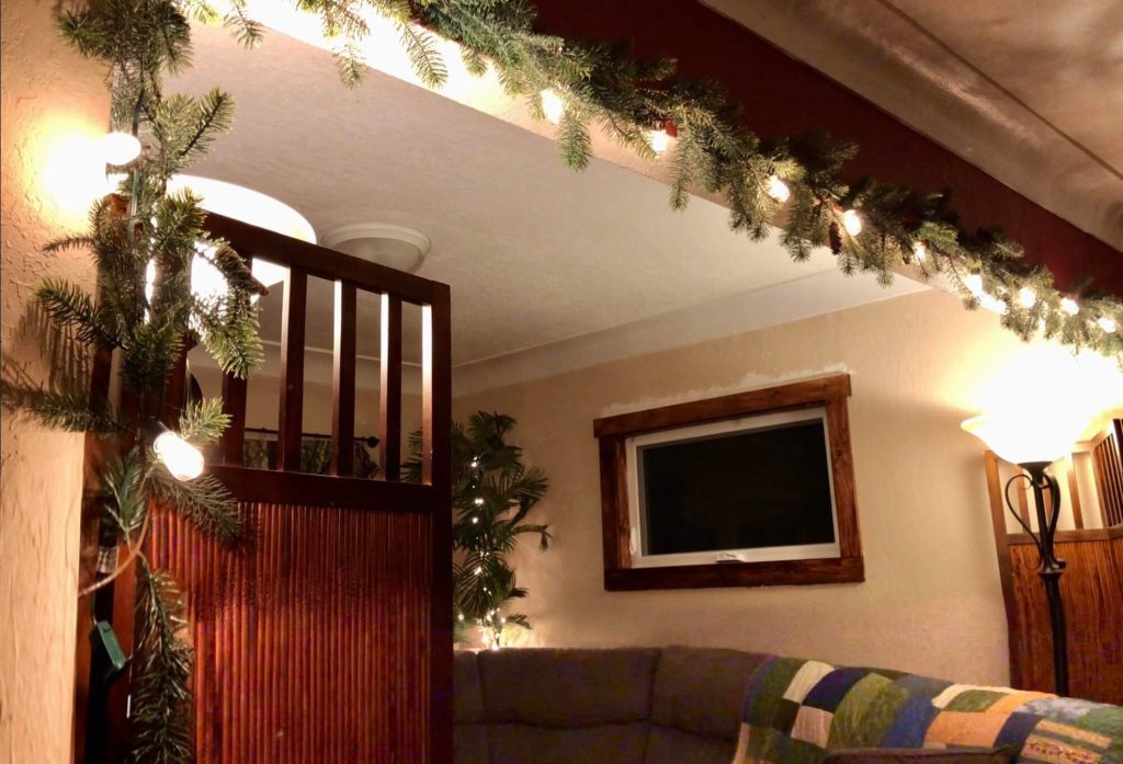 Photo of a room with string lights hanging with garland across a beam on ceiling. Two floor lamps are on, as are string lights around a plant in the corner of the room.