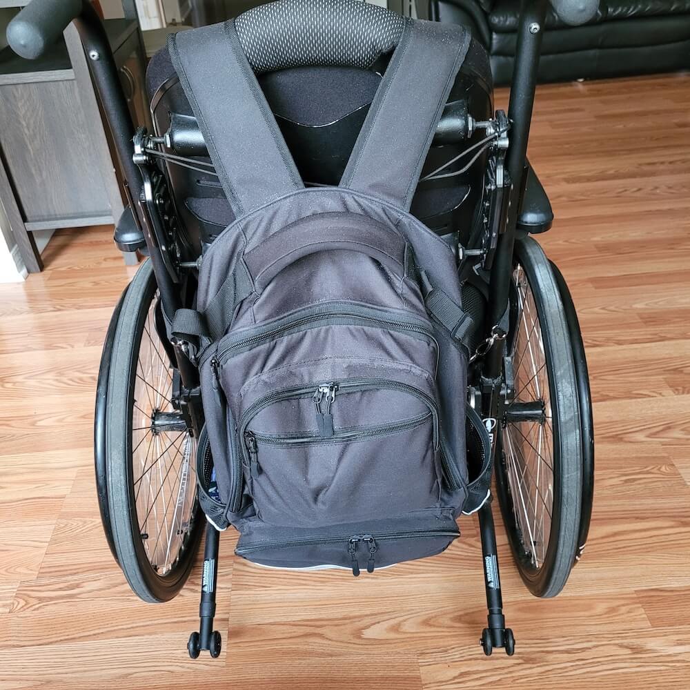 Black wheelchair bag hangs by two straps on the back of a wheelchair. The bag features several pockets with zippers and elastic pouches (for example, for bottles) on either side. There is a handle on top of the bag.
