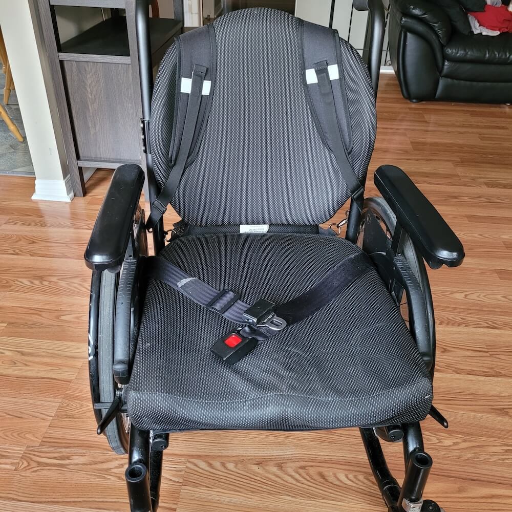 View of the straps holding the bag on the wheelchair, from the front of the wheelchair. The straps are on the back of the chair like a backpack.