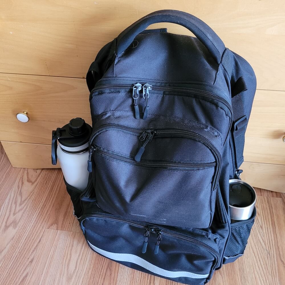 The backpack is sitting on the ground. A large water bottle and a travel coffee mug are in the two side pockets on the bag. A silver reflector strip is visible across the front pocket.