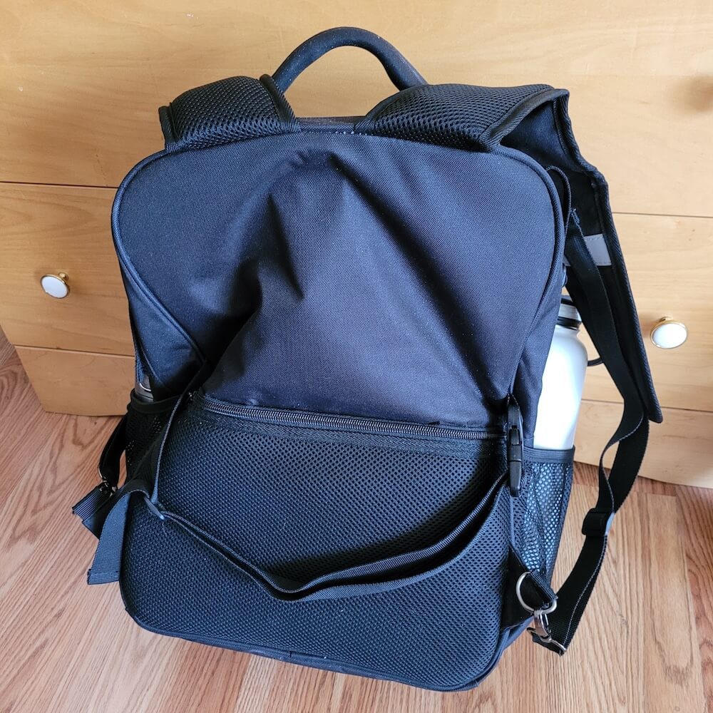 View of the back of the backpack with the straps lifted, revealing another strap that goes horizontally across the bag, as well as a "hidden" pocket, for which the zipper is half way up the back of the bag.