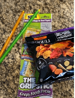 An orange and green Grip Stick are on a counter beside the packaging and a chip bag that has a purple Grip Stick across the top.