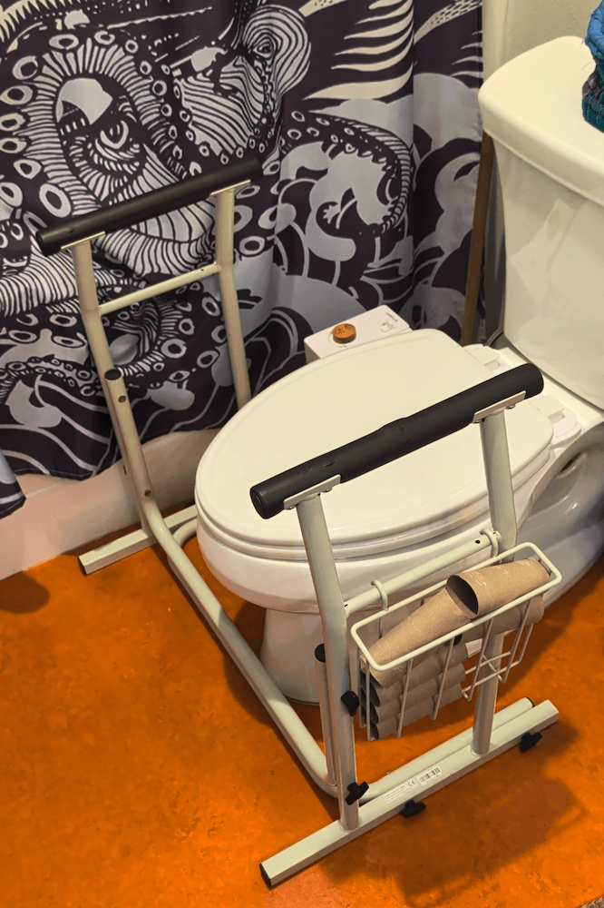 A frame with handles and a hanging storage basket filled with empty toilet roll tubes. The frame is positioned next to a toilet to aid with getting on and off.