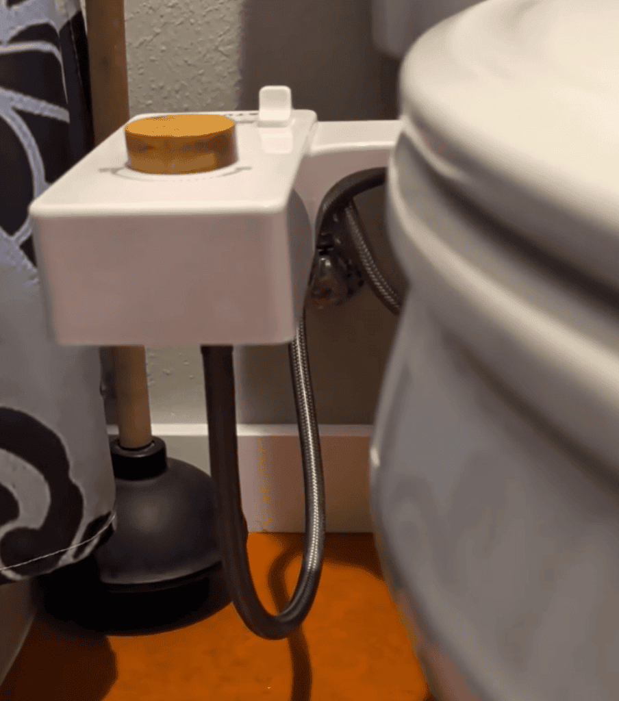 A toilet seat bidet. It's a unit attached to the side of a toilet with a dial and a metal hose attachment.
